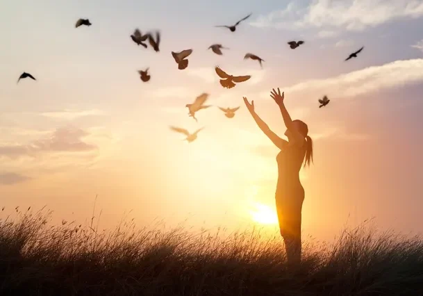 A woman standing in the grass with birds flying overhead.